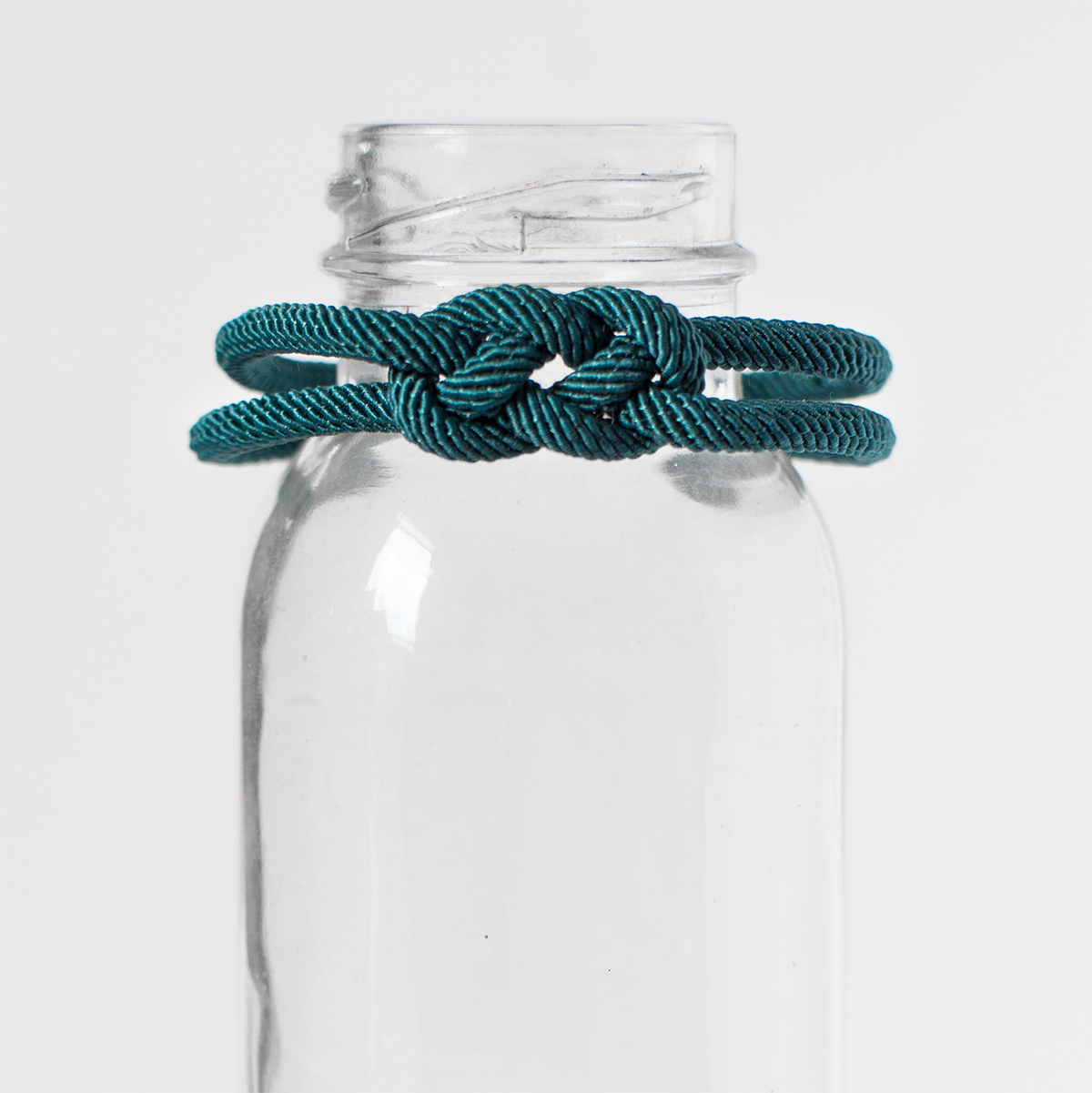Teal rope knot bracelet displayed on clear glass bottle, minimalist product photo